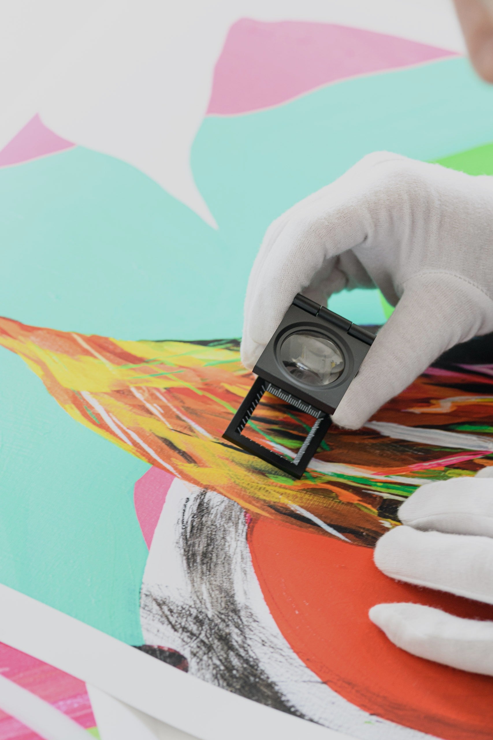 Gloved hand using loupe to inspect details of a colorful fine art print edition during quality control.