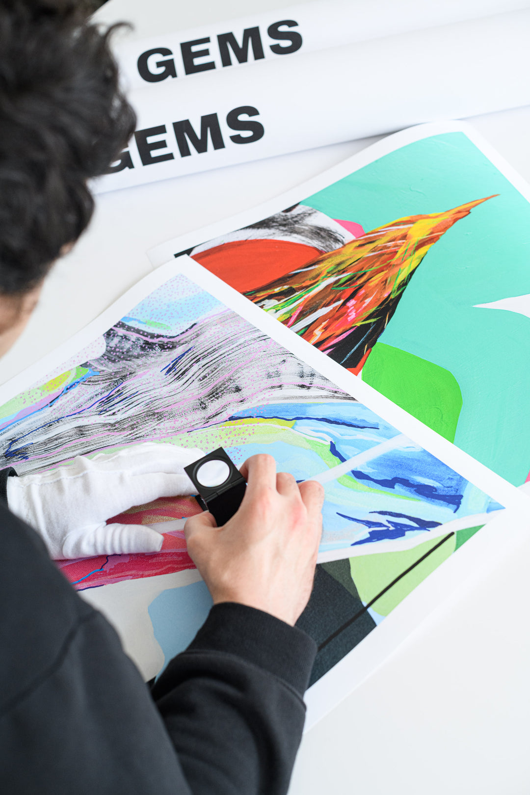 A person inspects a colorful abstract print with a linen tester while wearing white gloves, with two rolled GEMS art print tubes visible in the backgroun